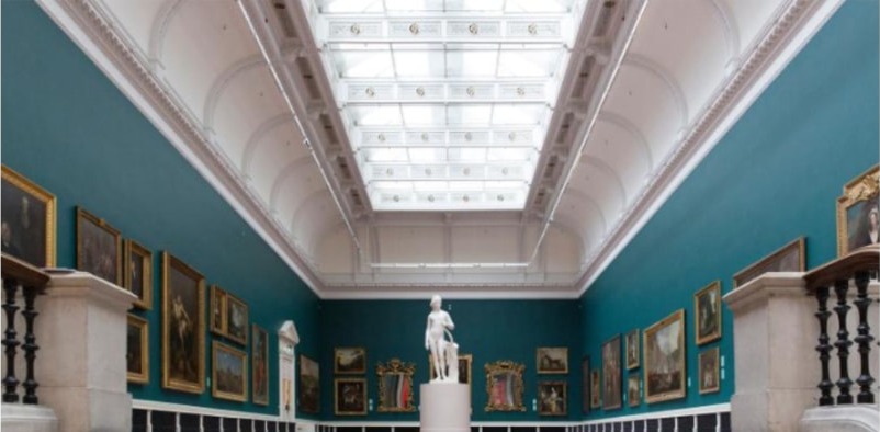 Interior of the National Gallery of Ireland — a teal-walled room of salon-hung paintings beneath a vaulted skylit ceiling, with a white sculpture on a pedestal.