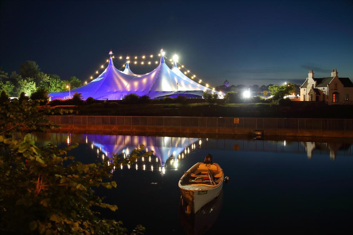 The Heineken Big Top at Fisheries Field lit up blue and white at night during Galway International Arts Festival, reflected in the river with a wooden rowing boat in the foreground