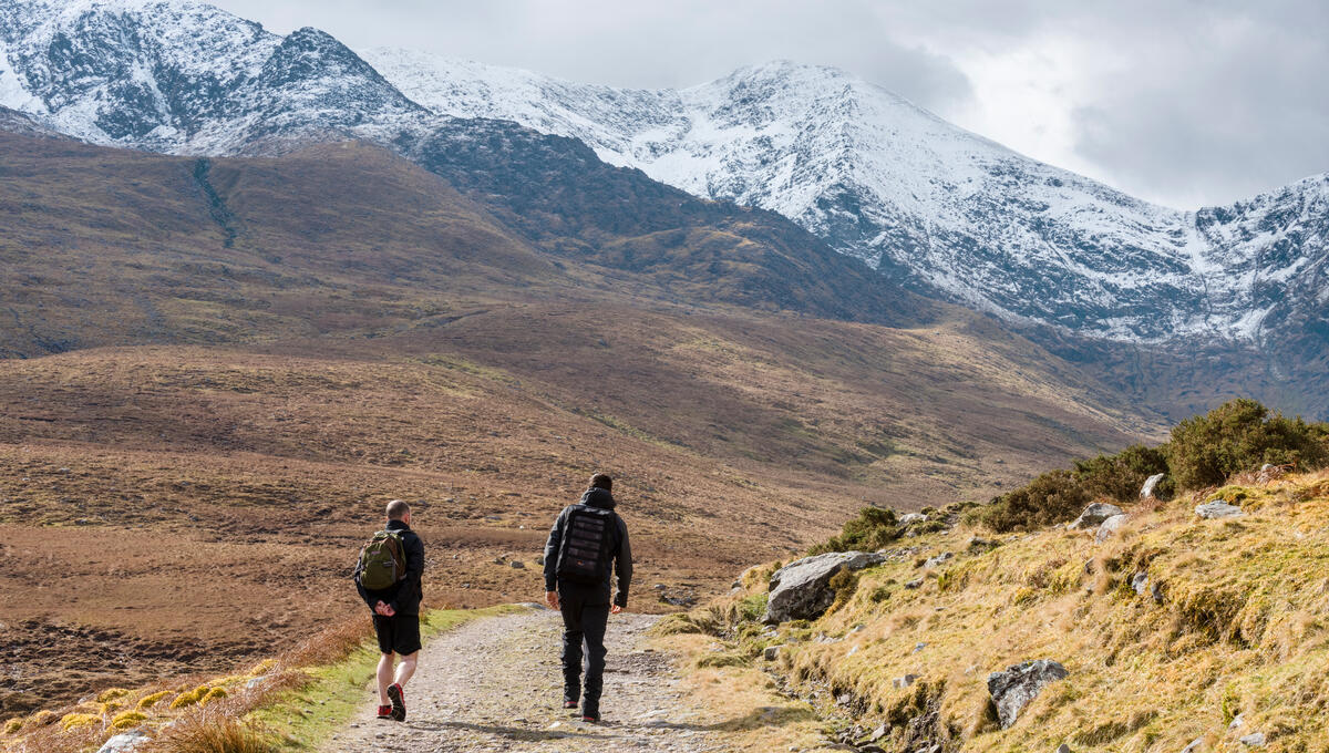 Two hikers on a mountain trail with snow-capped peaks rising in the distance