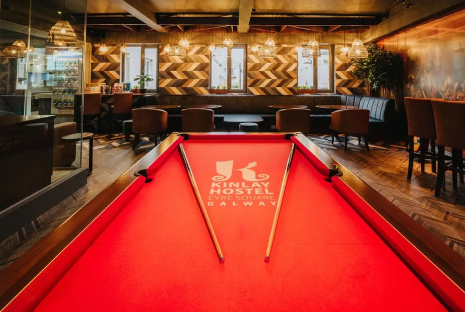 The Kinlay House common room on Eyre Square in Galway, with a red pool table in the foreground, leather armchairs, woven pendant lamps and a chevron wood accent wall