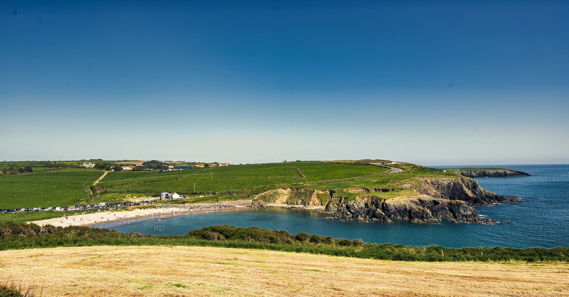 Kilmurrin Cove on the Copper Coast in County Waterford, with green cliffs, a sandy beach and clear blue water under a summer sky