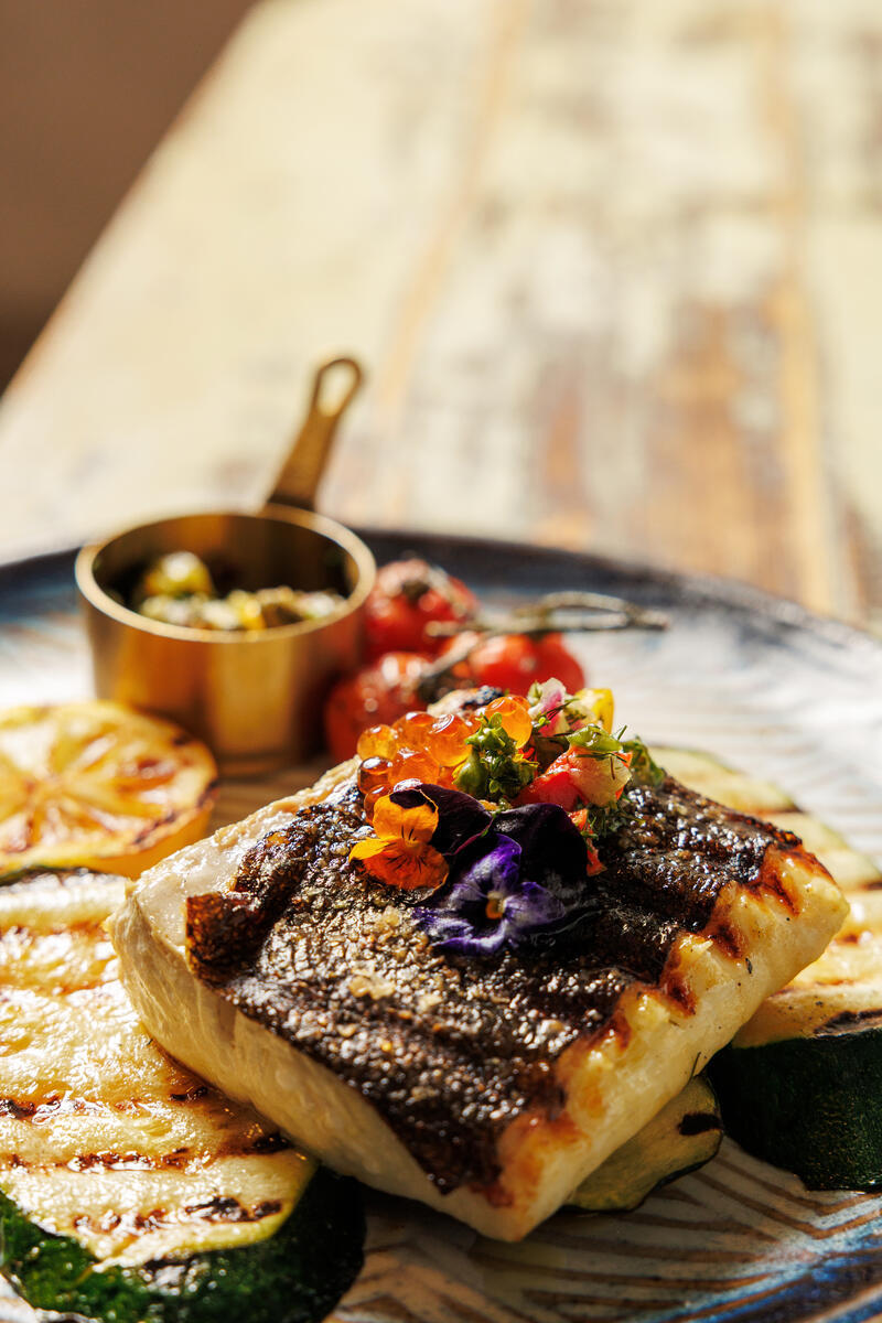 Pan-seared fish with grilled lemon, courgette, blistered tomatoes and edible flowers on a ceramic plate